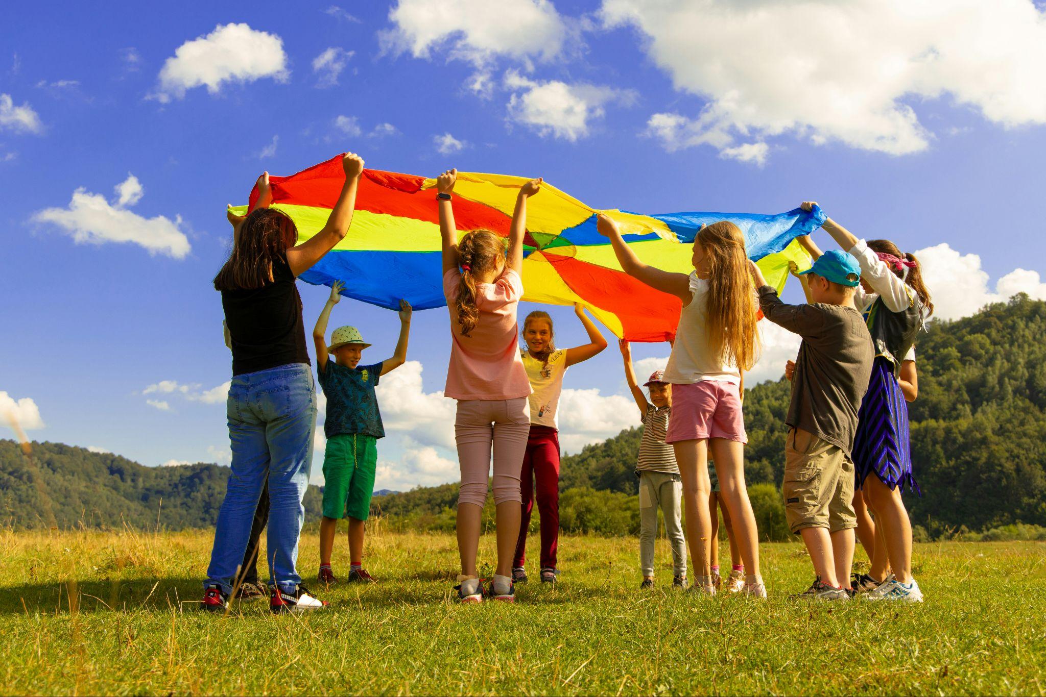 Children standing in a grassy outdoor setting holding a colorful parachute overhead, symbolizing community, childhood, and collective protection